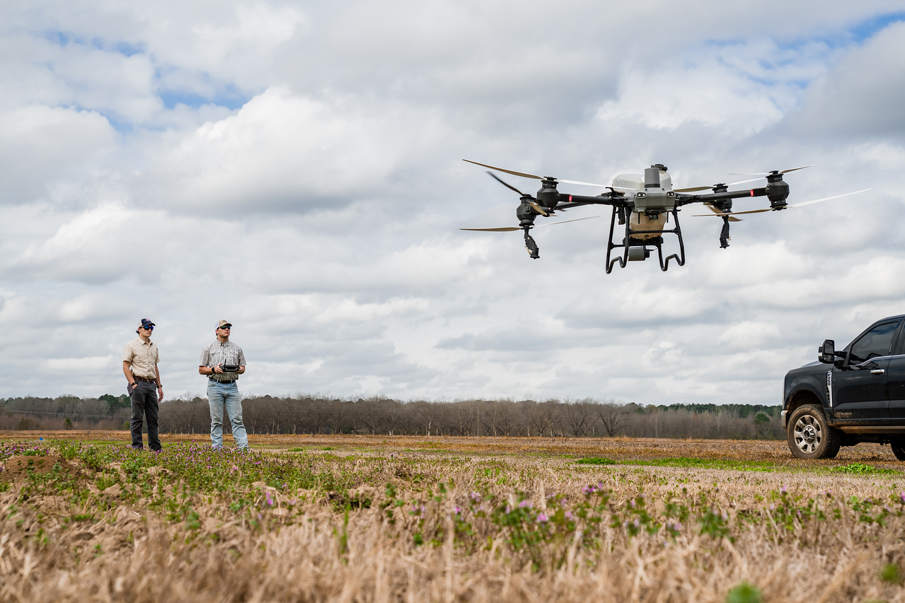 Spray drone in flight over a row-crop field with the founders at the controls